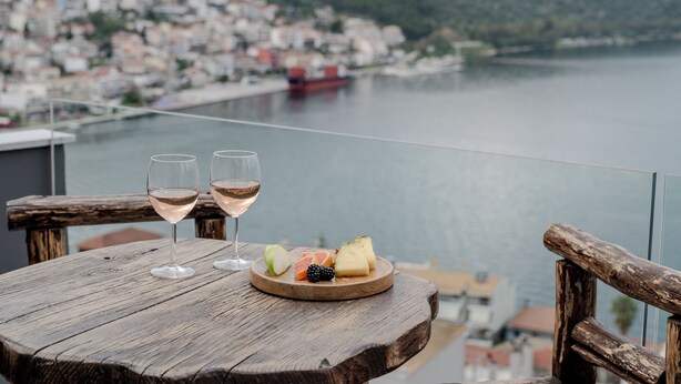 A wooden table with two glasses of white wine and a plate of fruit on a balcony overlooking a body of water and a town.