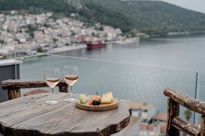 Ein Holztisch mit zwei Gläsern Weißwein und einer Obstplatte auf einem Balkon mit Blick auf ein Gewässer und eine Stadt.