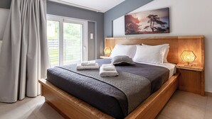 A neatly made bed with a dark bedspread, two pillows, and folded towels on a wooden platform. There is a nightstand with a lamp and a painting of a tree on the wall above the bed.