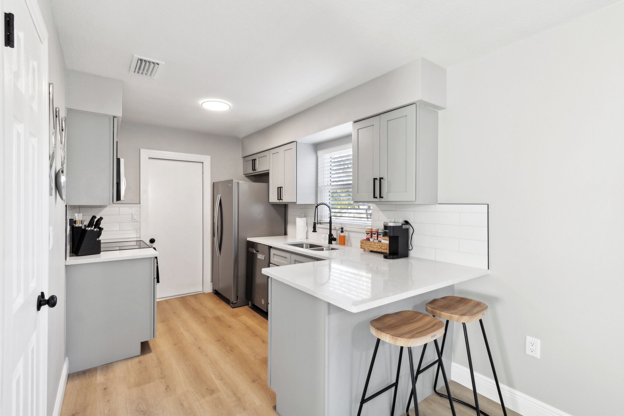 Una cocina moderna con armarios grises, un refrigerador de acero inoxidable, una encimera blanca y dos taburetes de madera.