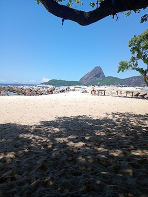 Ein Strand mit felsigem Ufer, ein Berg im Hintergrund und ein Baum, der Schatten spendet.