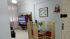 Un comedor con una mesa de vidrio, sillas de madera, un televisor sobre un mueble bajo y un cuadro en la pared.