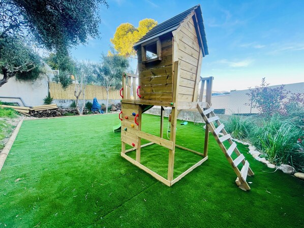 A wooden playhouse with a slide and climbing wall, set on a green lawn with a fence and trees in the background.