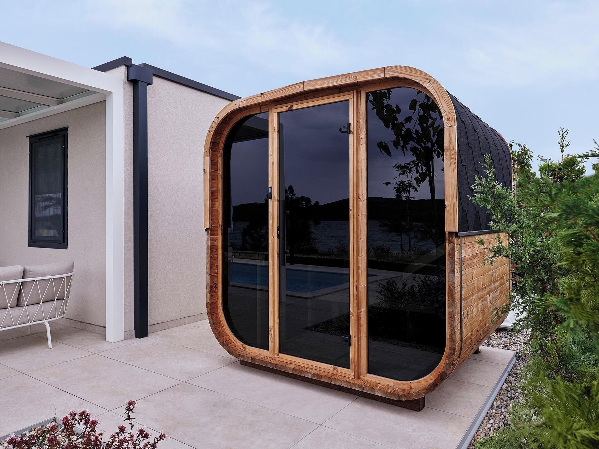 Une pièce moderne, avec une structure en bois et des parois vitrées, dotée d’une porte coulissante, et située à côté d’une chaise blanche et d’une porte vitrée donnant sur une piscine.