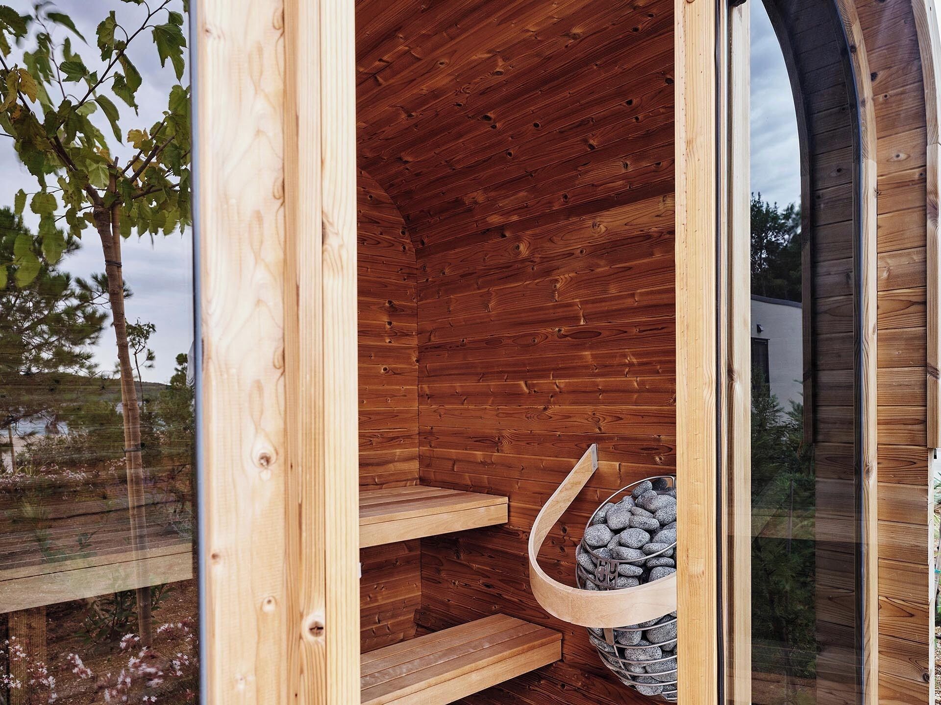 A wooden sauna with a glass door, a wooden bench, and a basket of stones.