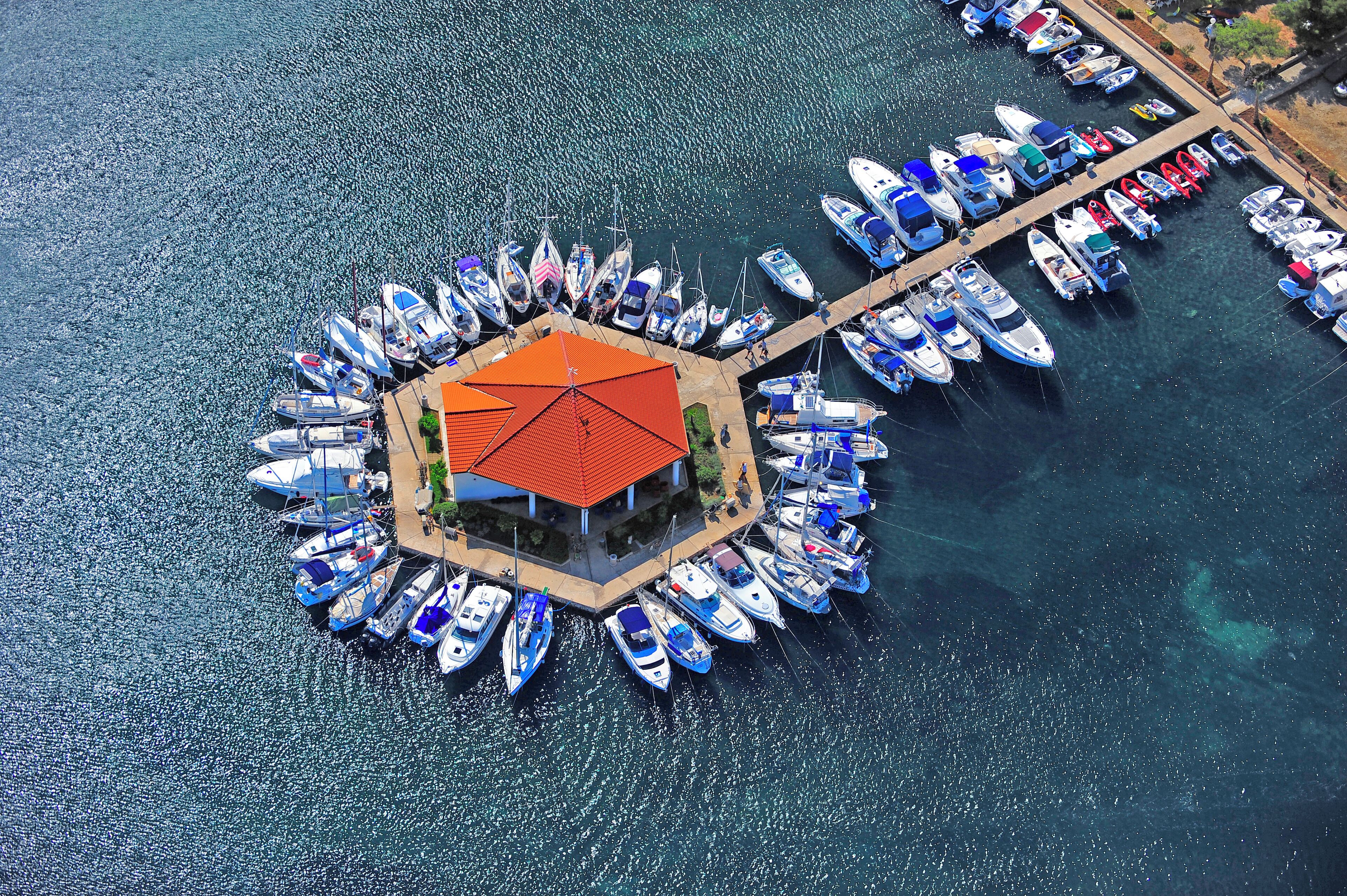 An aerial view of a marina with numerous boats docked around a central island with a red-roofed structure.