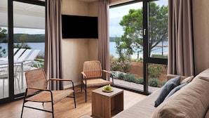 Een moderne woonkamer met een bank, twee rieten fauteuils, een salontafel en een aan de muur bevestigde televisie. Grote ramen bieden uitzicht op een meer en groen.