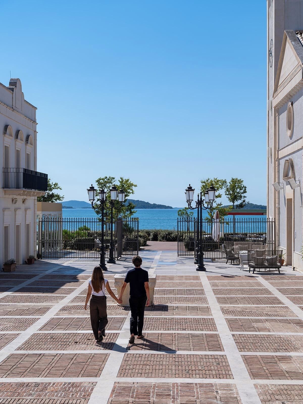 A couple walking on a paved pathway towards a scenic view of the sea, with buildings and street lamps on either side.