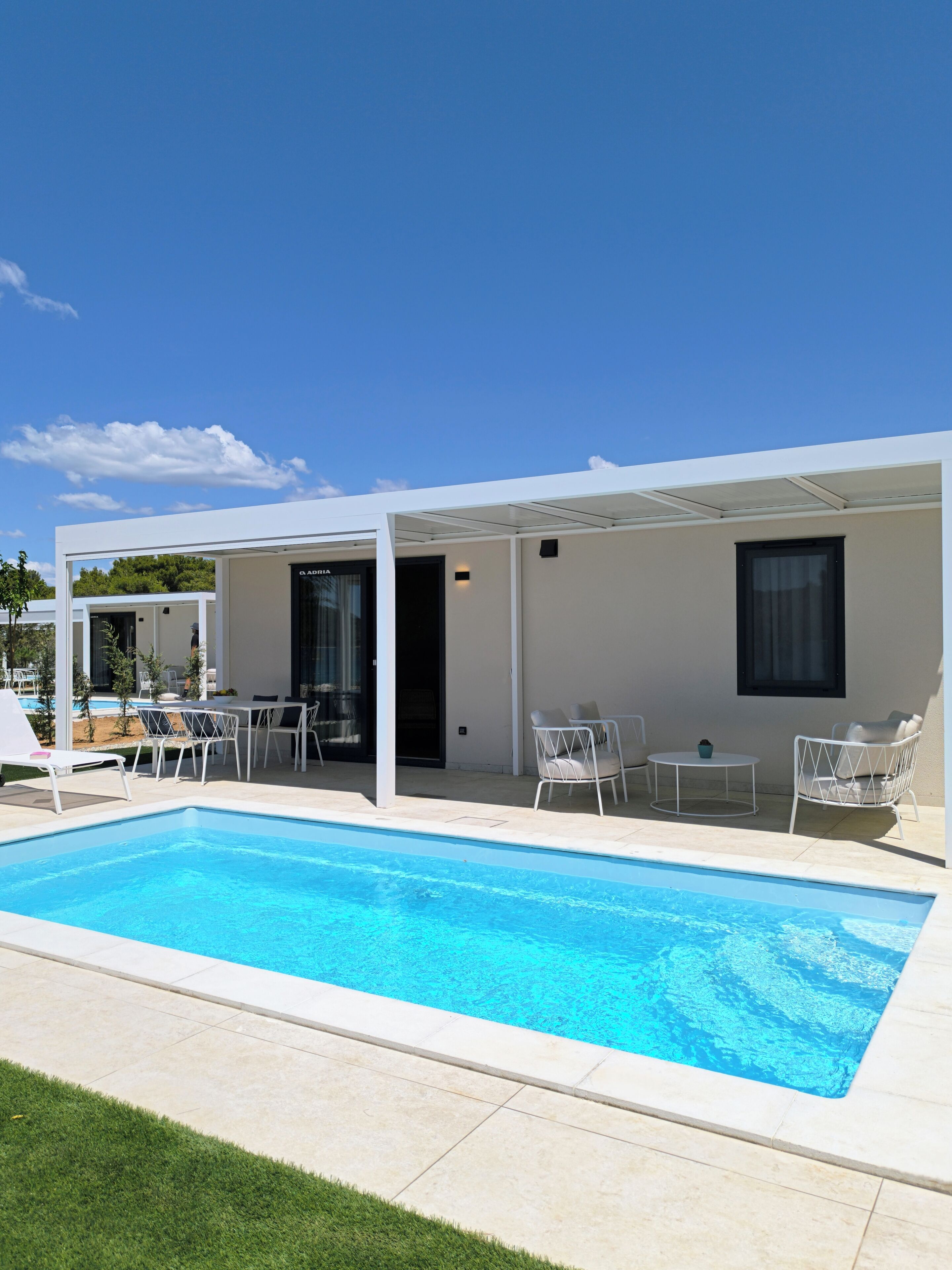 Un espace moderne aménagé au bord de la piscine, avec une piscine à l’eau d’un bleu limpide, des meubles d’extérieur blancs et un coin salon abrité.