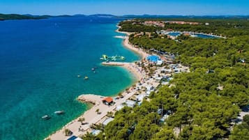 Vue aérienne d’une zone côtière avec une plage, des eaux d’un bleu limpide et une zone densément boisée où se trouvent des bâtiments.