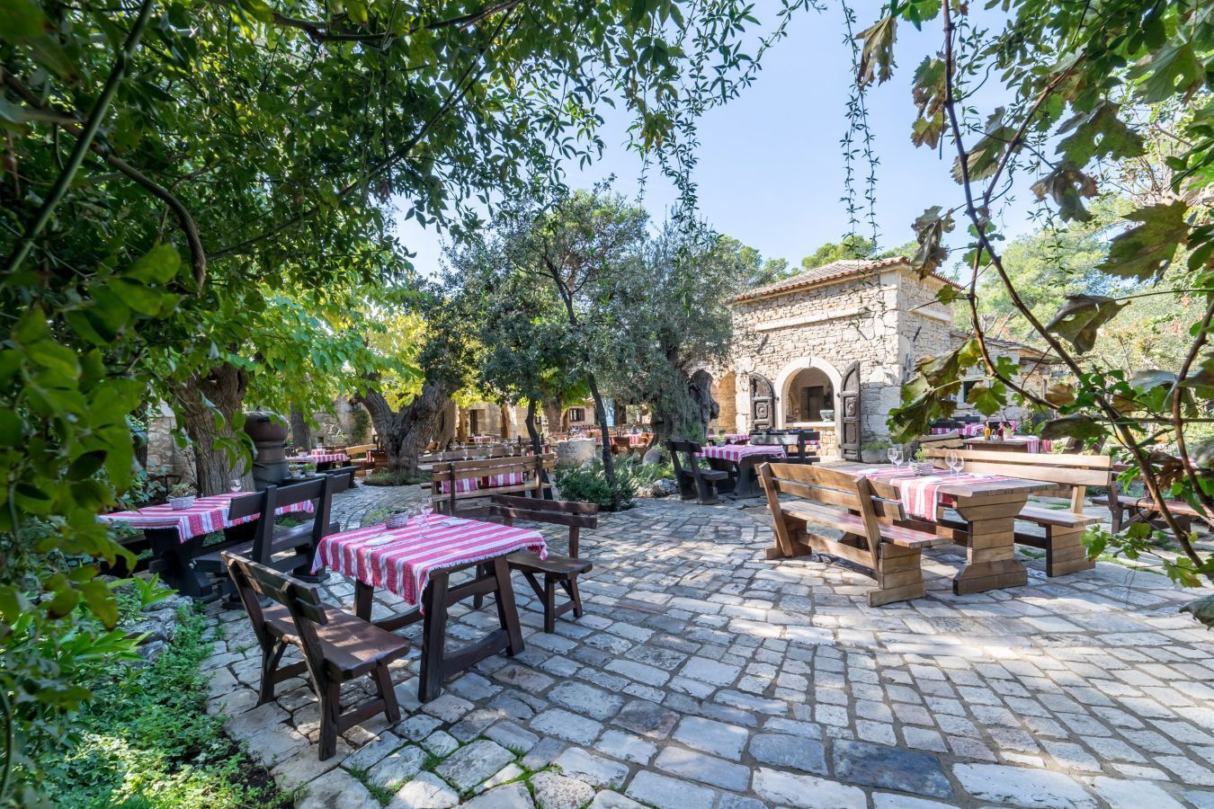 An outdoor dining area with stone tables and benches, surrounded by trees and a stone building with arches.