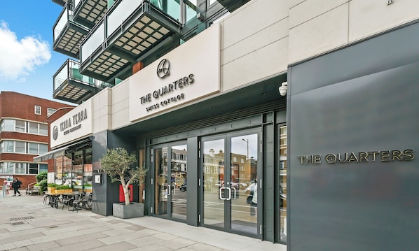 Un edificio moderno con el letrero "The Quarters", que cuenta con una entrada de vidrio y una jardinera con un árbol en el exterior.