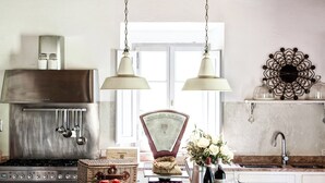 A kitchen with a gas stove, oven, hanging pots, a scale, a bowl of tomatoes, a wine bottle, and a vase of flowers.