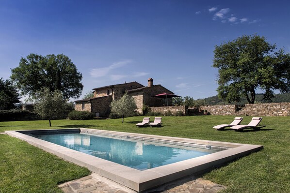 Una piscina rectangular con dos sillones de descanso sobre el césped, un edificio de piedra y árboles al fondo.