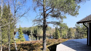 A wooden deck overlooking a lake with trees and a small building in the background. - Holiday home Tived - directly on lake, sauna, boat, hot tub (Tivedstorp)