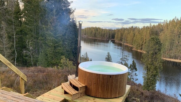 A hot tub on a wooden deck with a view of a lake and forest. - Holiday home Tived - directly on lake, sauna, boat, hot tub (Tivedstorp)