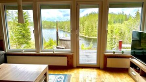 A room with large windows offering a view of a lake and forest. There is a wooden table, a TV, and a radiator. - Holiday home Tived - directly on lake, sauna, boat, hot tub (Tivedstorp)