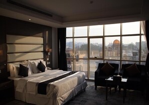 A hotel room with a large bed, a desk, two chairs, and a large window offering a city view. - Asal Al Rimal Hotel For Hotel Service (Jeddah)