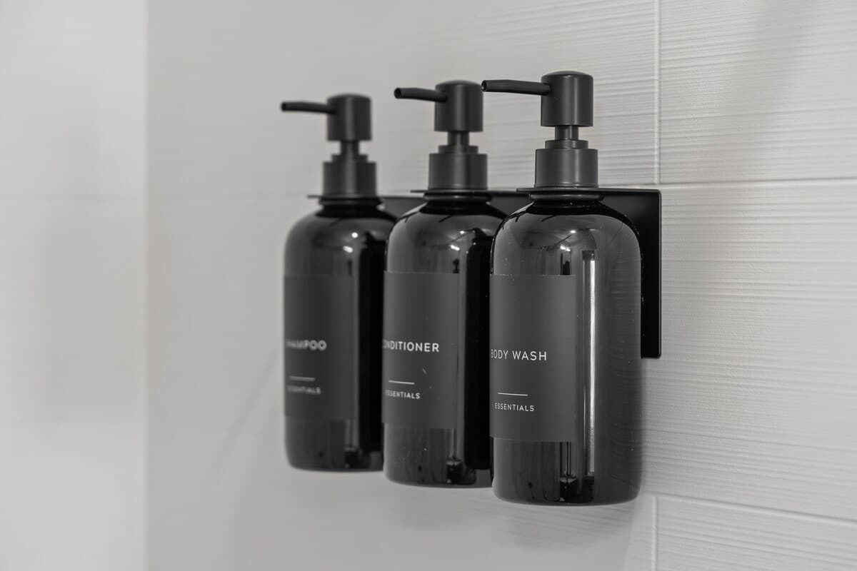 Tres botellas negras de champú, acondicionador y gel de baño sujetas a una pared de baldosas blancas.