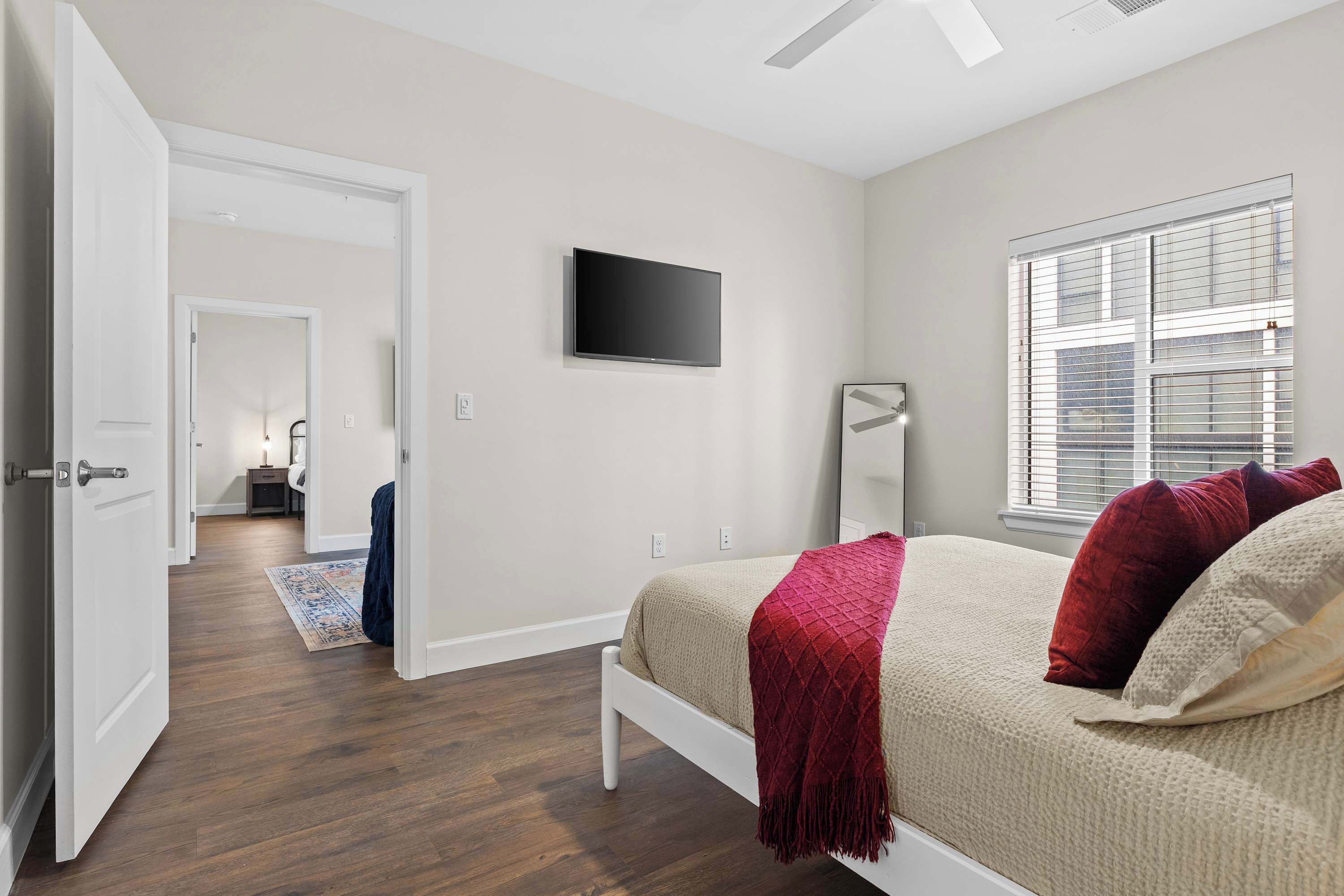 A bedroom with a bed, a TV mounted on the wall, a window with blinds, and a door leading to another room.
