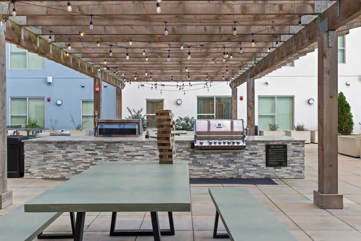 An outdoor patio with a stone barbecue station, a wooden pergola with string lights, and a long table.