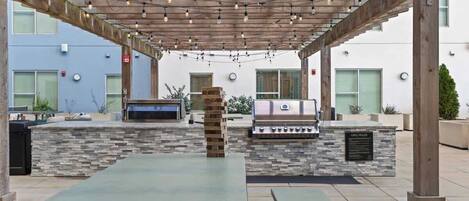 An outdoor patio with a stone barbecue station, a wooden pergola with string lights, and a long table.