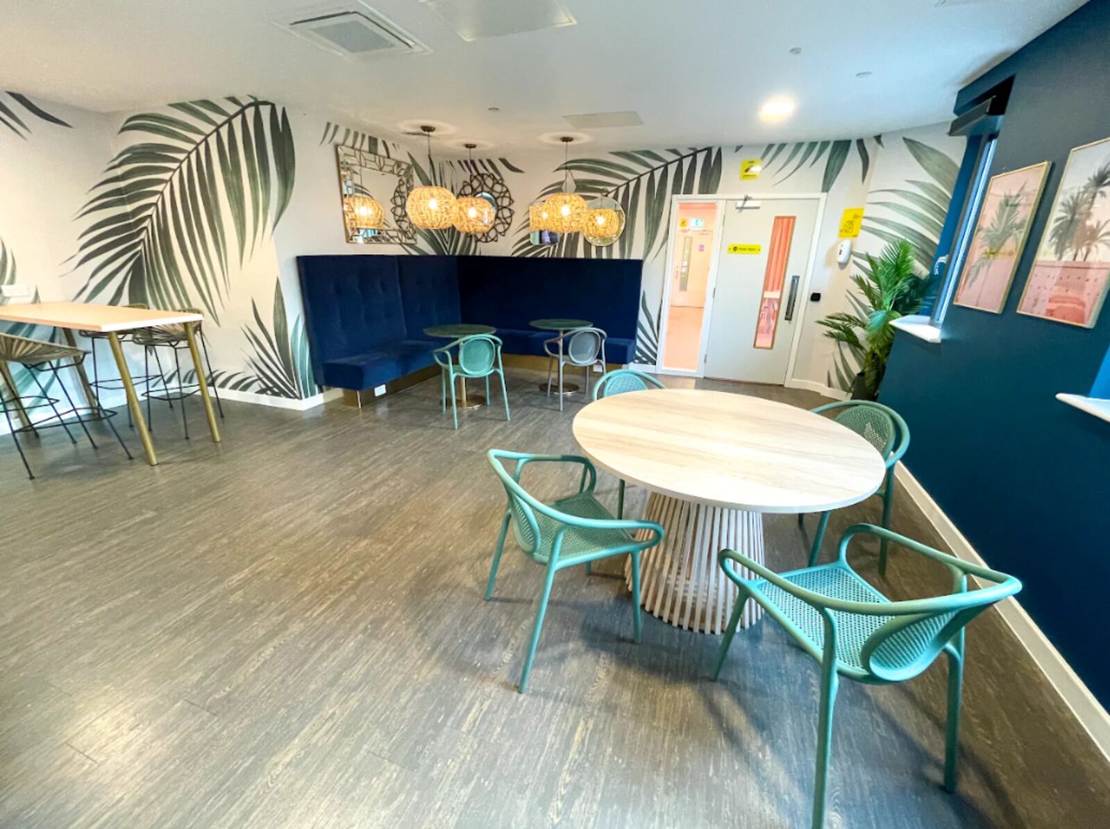 A modern cafe with a wooden floor, teal chairs, a round table, and a blue accent wall with palm leaf designs.