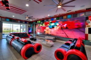 A modern lounge area with a large TV screen displaying a festive animation, ceiling fans, and a seating area with red and black cushions.