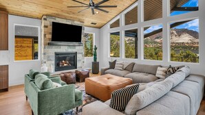 A spacious living room with a fireplace, large windows, and a ceiling fan. - High Desert Home | Modern Luxury Near Zion NP (Orderville)