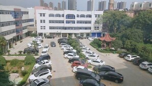 A parking lot with multiple cars, a building with a modern facade, and a traditional-style structure with a red roof. - 7Days Inn Hotel (Zhoukou Wuyi Square City Central Hospital) (Zhoukou)