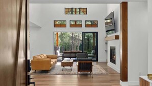 A modern living room with a sofa, coffee table, and armchair, a fireplace, and large windows with a view of trees.