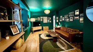 Un salon chaleureux avec un canapé en cuir marron, une table basse en bois et un tapis vert. Des tableaux encadrés ornent les murs, et une lampe est posée sur une table d’appoint.