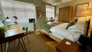 Une chambre à coucher comprenant un lit, une table de chevet, un bureau avec une chaise, un radiateur, une fenêtre avec des stores et une lampe fixée au mur.
