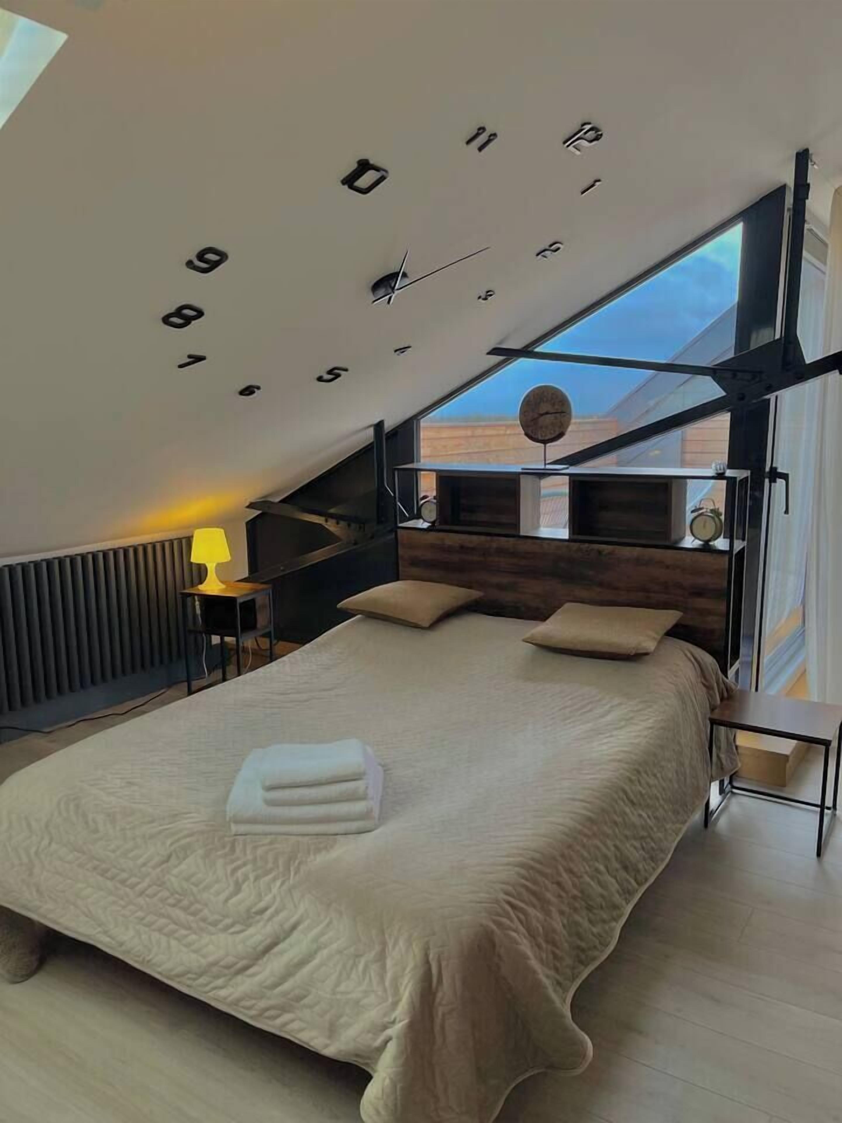 Une chambre moderne avec un plafond en pente, un grand lit, une table de chevet, une lampe et une vue sur l’extérieur.