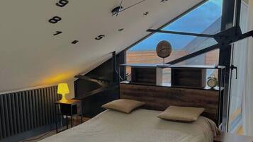 Une chambre moderne avec un plafond en pente, un grand lit, une table de chevet, une lampe et une vue sur l’extérieur.