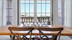 A dining area with a wooden table and chairs, large windows with blinds, and a view of buildings outside.