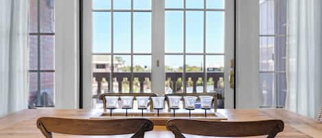A dining area with a wooden table and chairs, large windows with blinds, and a view of buildings outside.