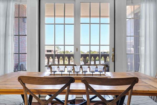 A dining area with a wooden table and chairs, large windows with blinds, and a view of buildings outside. - The Village Blue Haven Condo - Private Beach Access, Pool & Hot Tub! (Rosemary Beach)