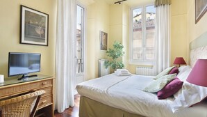 A hotel room with a bed, a desk with a computer, a chair, a window with curtains, a radiator, and a plant.