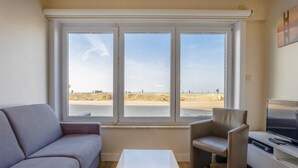 A living room with a sofa, a chair, a small table, and a TV. There are three large windows with a beach view. - Renovated Ground-floor Apartment in Koksijde (Koksijde)
