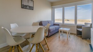 A modern living room with a white dining table, white chairs, a gray sofa, and a small white side table. There is a large window with a view of the beach. - Renovated Ground-floor Apartment in Koksijde (Koksijde)