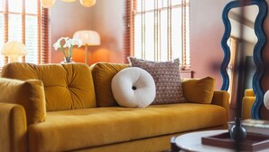 Un canapé de couleur moutarde agrémenté de coussins à motifs, une petite table d’appoint ronde et un miroir décoratif à cadre ondulé.