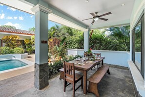 An outdoor patio with a pool, wooden table, and chairs, surrounded by lush greenery and a clear blue sky. - Stylish Home with Chic Decor, Luxury Amenities & Tropical Outdoor Oasis with Sparkling Private Pool (Cabo Velas)