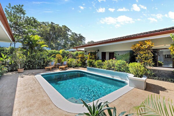 A swimming pool with lounge chairs, surrounded by lush greenery and a building with a tiled roof. - Stylish Home with Chic Decor, Luxury Amenities & Tropical Outdoor Oasis with Sparkling Private Pool (Cabo Velas)