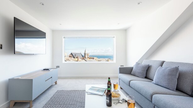 A modern living room with a large window, a grey sofa, a coffee table with a bottle and glasses, and a TV mounted on the wall.