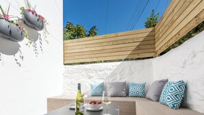 An outdoor seating area with a wooden bench, cushions, and a table with a bottle and glass.