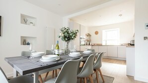 A modern dining room with a long table set for six, a kitchen in the background, and a large window providing natural light.