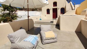 Área externa ensolarada com dois cadeirões, um guarda-sol branco e um edifício com porta vermelha e janelas em arco.