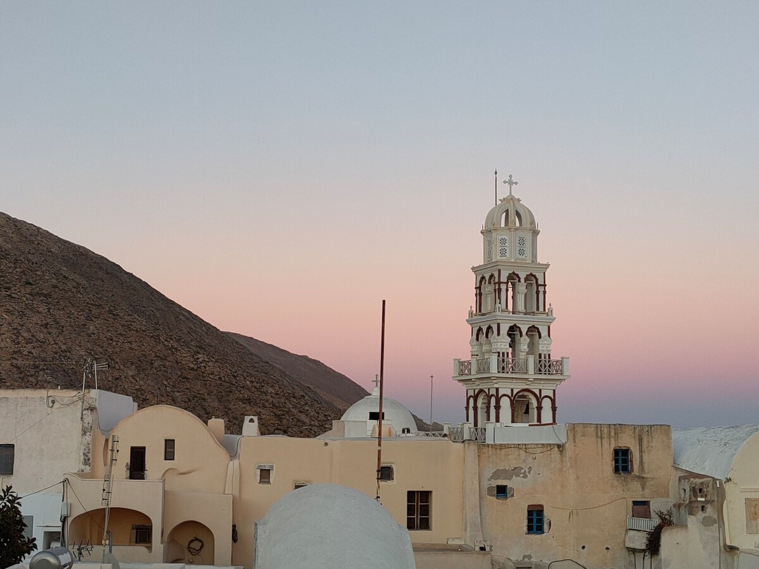 Uma igreja com torre sineira e cúpula, rodeada por edifícios com janelas em arco e uma colina ao fundo.