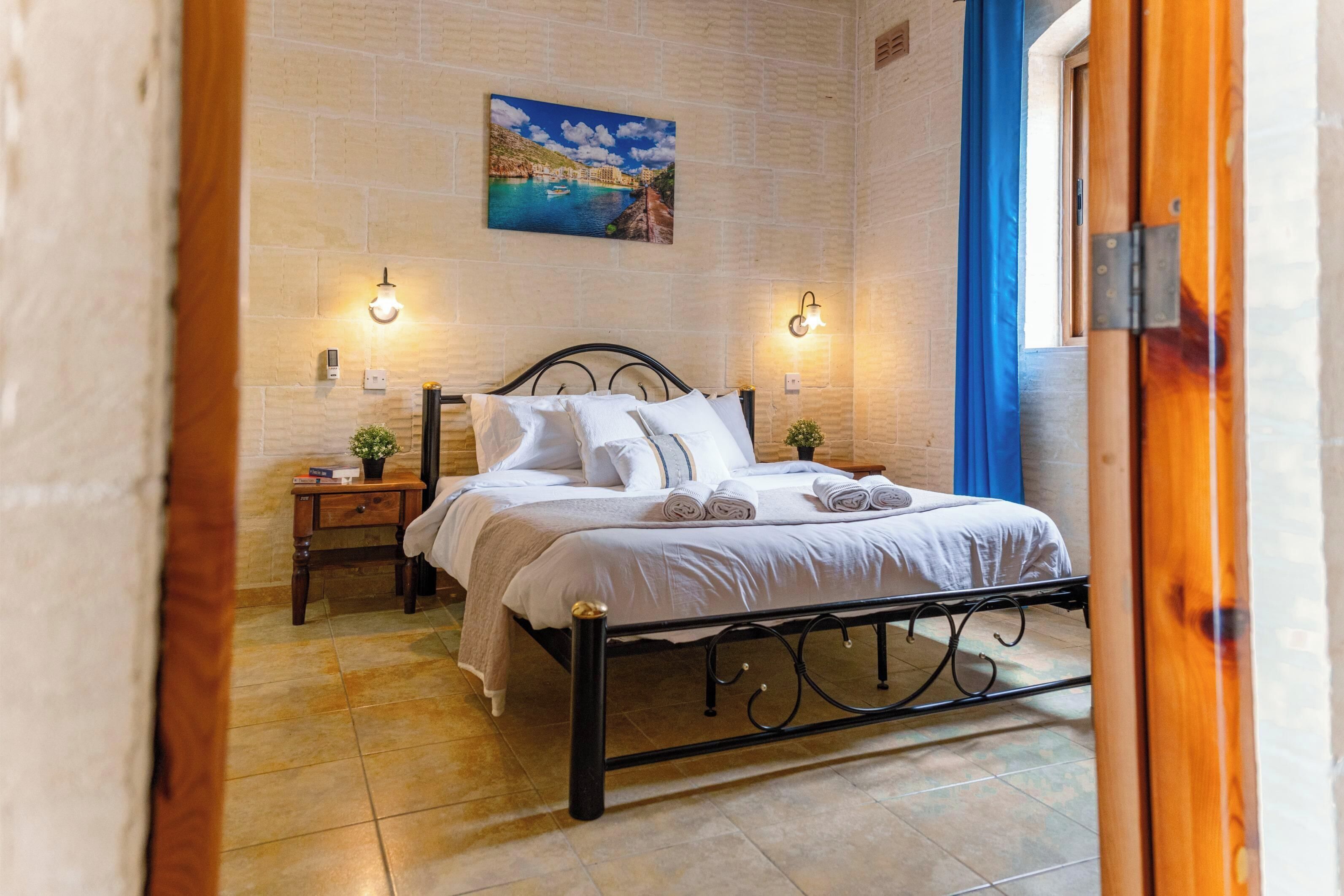 Una cama bien hecha con ropa de cama blanca, dos mesitas de noche con lámparas y un cuadro de una escena costera en la pared.
